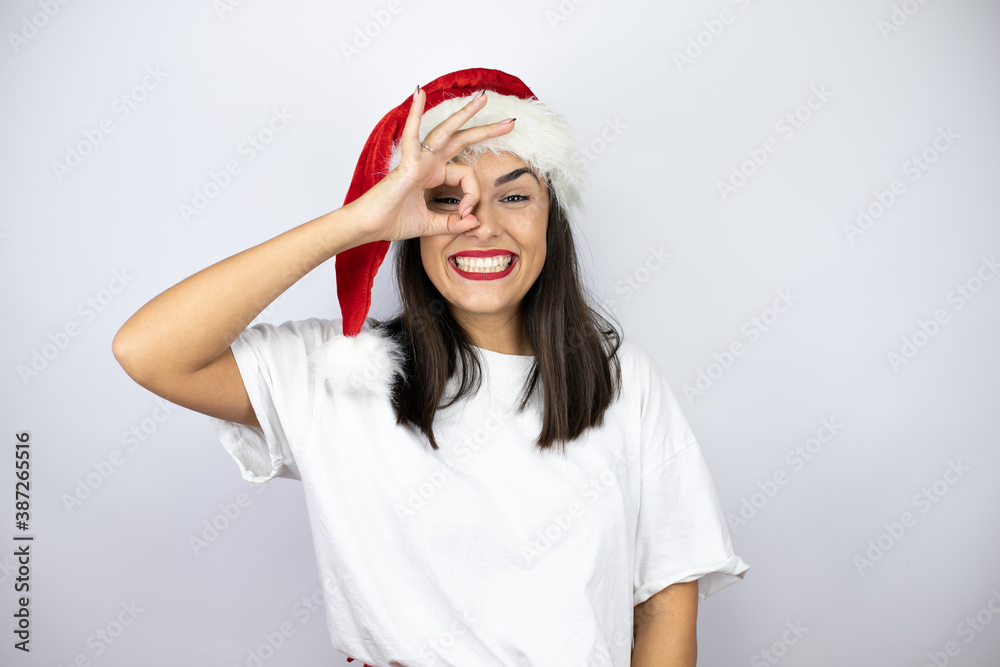 Young beautiful woman wearing a christmas hat over white background doing ok gesture shocked with smiling face, eye looking through fingers