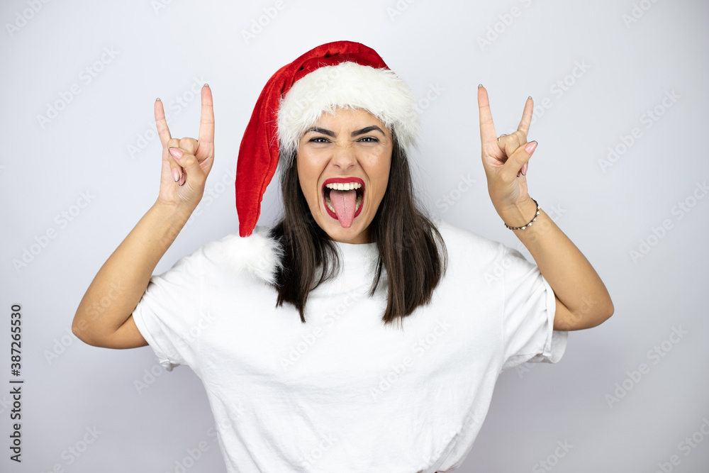 Young beautiful woman wearing a christmas hat over white background shouting with crazy expression doing rock symbol with hands up Young beautiful woman wearing a christmas hat over white background shouting with crazy expression doing rock symbol with hands up