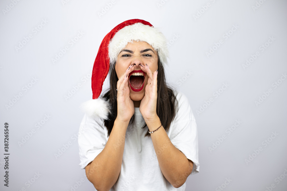 Young beautiful woman wearing a christmas hat over white background shouting and screaming loud to side with hands on mouth Young beautiful woman wearing a christmas hat over white background shouting and screaming loud to side with hands on mouth