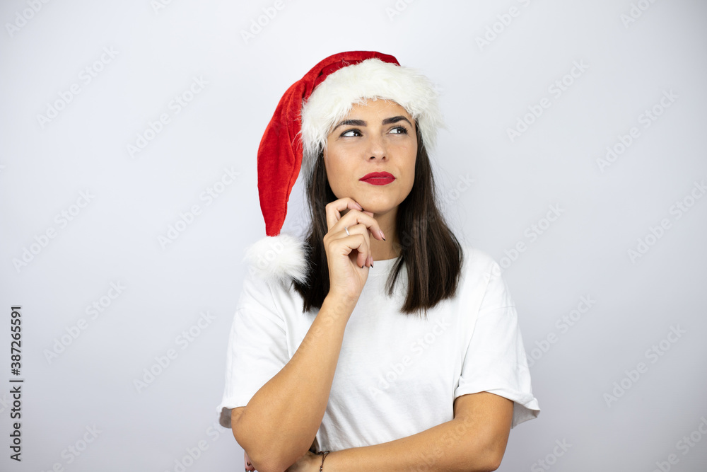 Young beautiful woman wearing a christmas hat over white background thinking and looking to the side Young beautiful woman wearing a christmas hat over white background thinking and looking to the side