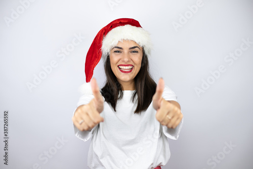 Young beautiful woman wearing a christmas hat over white background smiling and doing the ok signal with her thumbs