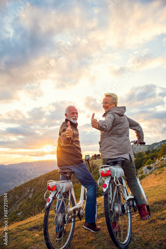 Elderly man and woman on e-bikes holding their thumbs up on mountain. Beautiful sunset at background.