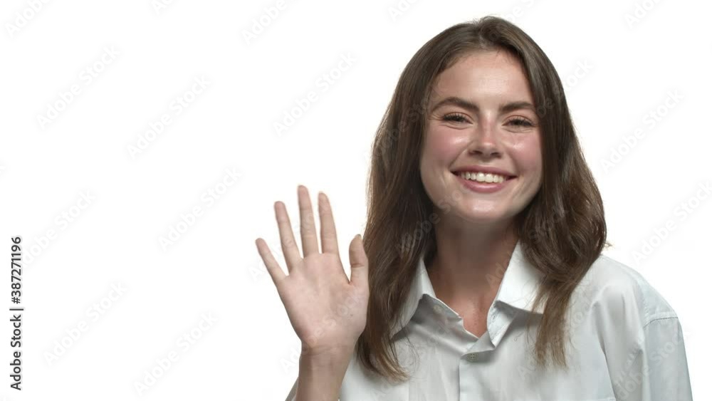 Close-up of attractive brunette woman saying hi and waving hand, smiling at camera, standing over white background Close-up of attractive brunette woman saying hi and waving hand, smiling at camera, standing over white background