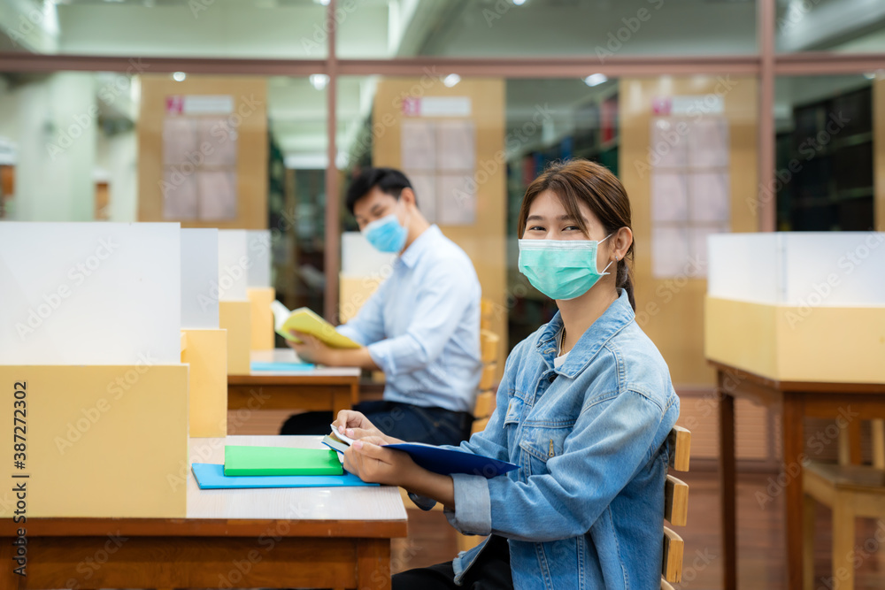 Two Asian university students wearing face mask and sitting and smile in library social distance from other 6 feets to avoid the spread of coronavirus in University.