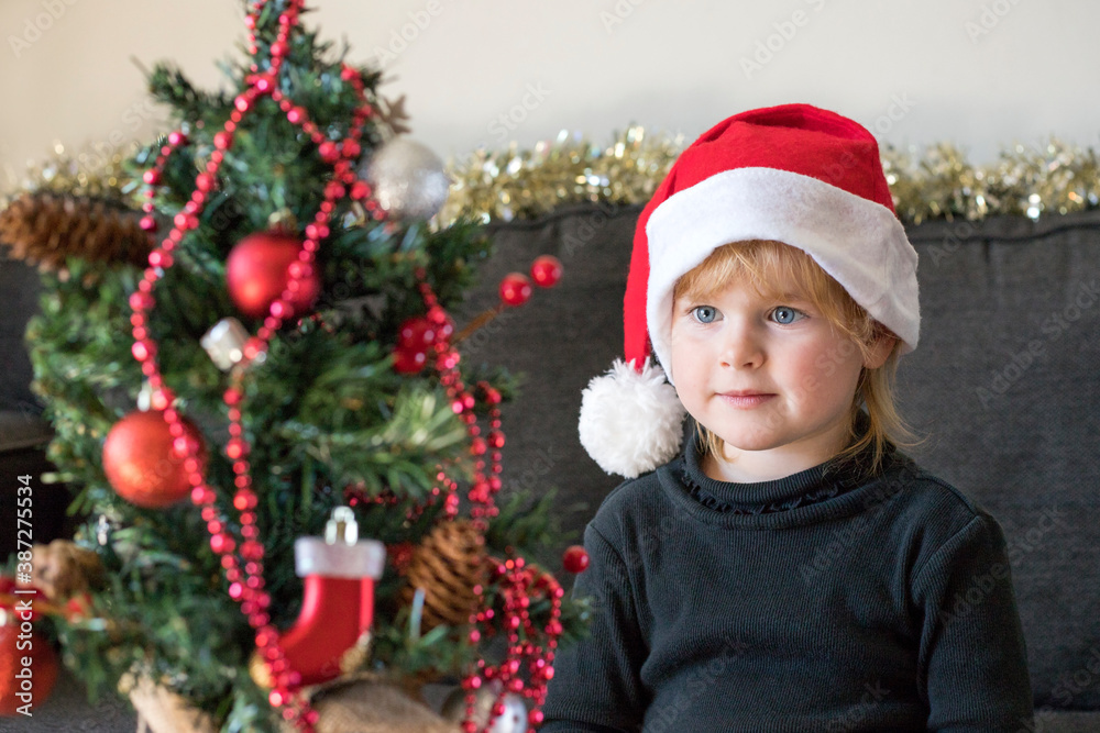 Christmas and New Year holidays preparation concept. Happy smiling child decorating  and looking at Christmas tree, wearing dress and santa hat. 