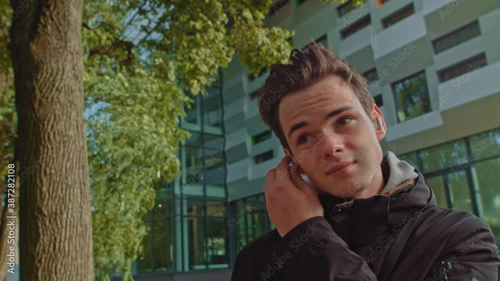 Young man talking on the phone using wireless headphones outdoors while walking down the street. young guy, teenager wears modern wireless headphones. Portrait of a young man.