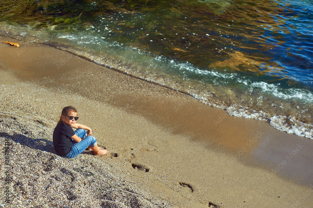 A child is resting on the seashore . A little girl sits on the sand on an autumn day at the sea . The view from the top . A child is resting on the seashore . A little girl sits on the sand on an autumn day at the sea . The view from the top .
