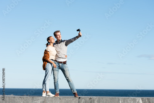Social media content creation concept. A young couple taking themselves a selfie with a compact camera in a sunny day at the seaside.