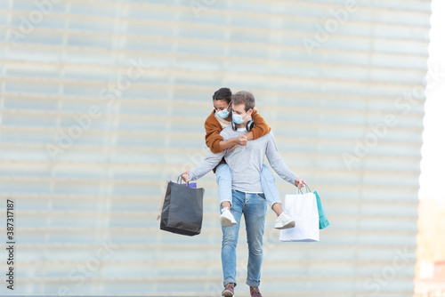 Shopping during Coronavirus pandemic concept. A young man carrying his girlfriend on the back while holding their shopping bags.