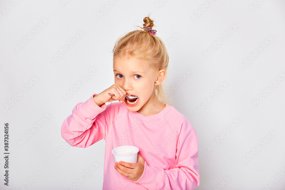 Cute little girl eating tasty yogurt on gray background. 4-5 year old kid posing in studio in a pink sweatshirt. Space for text Cute little girl eating tasty yogurt on gray background. 4-5 year old kid posing in studio in a pink sweatshirt. Space for text