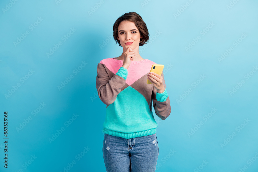 Photo portrait of curious woman holding phone in one hand touching chin with finger isolated on pastel light blue colored background Photo portrait of curious woman holding phone in one hand touching chin with finger isolated on pastel light blue colored background