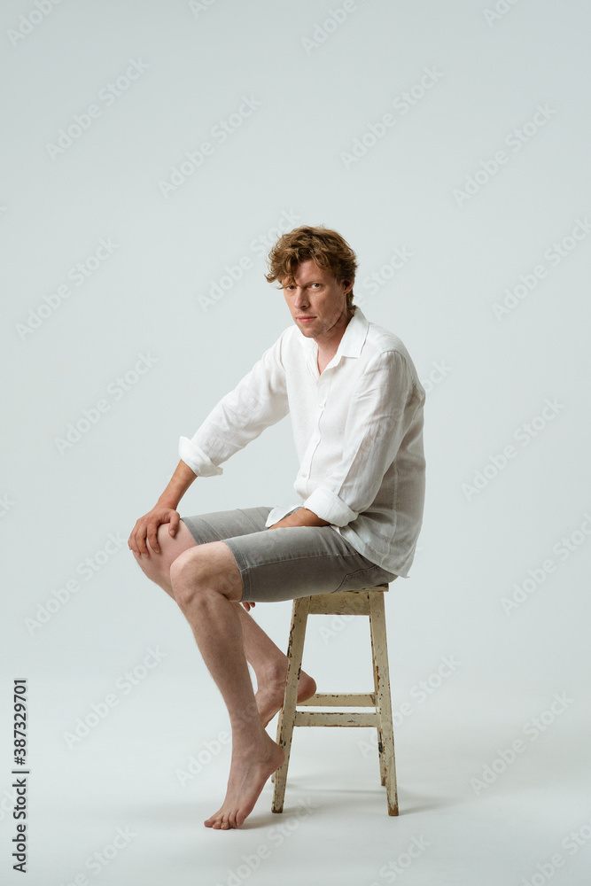 Young man sitting barefoot on tall stool in white studio. Full length picture. Isolated on white.