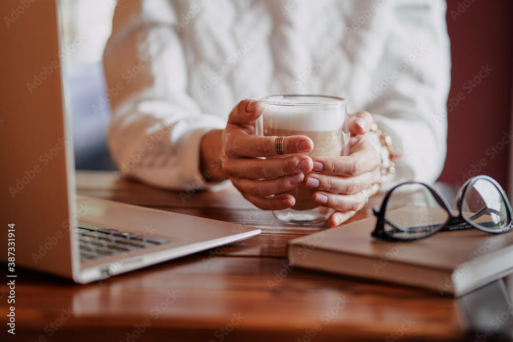 Working and relaxing at cafe. Close up of female hands holding cup of cappuccino coffee on wooden tavble with laptop computer.