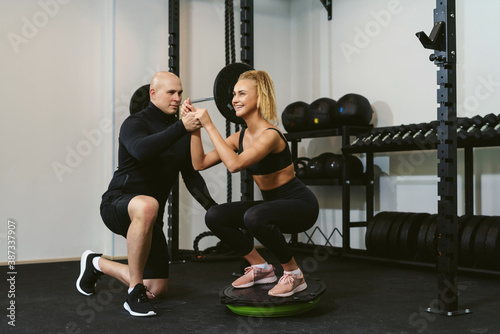 A young sportive woman trains butt and legs on a balance board performing lunges and squats. Handsome trainer assisting her and monitoring her sport technique. Personal training with a coach