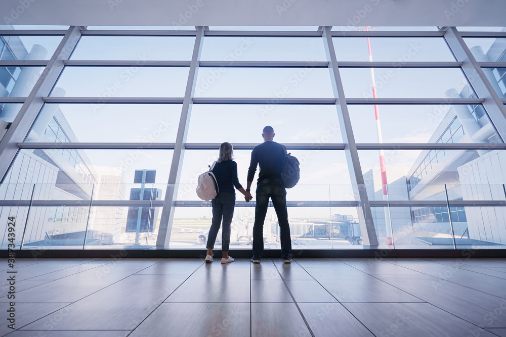 Traveling concept. Back view of loving couple in casual wear standing near the window of international airport terminal. Traveling concept. Back view of loving couple in casual wear standing near the window of international airport terminal.