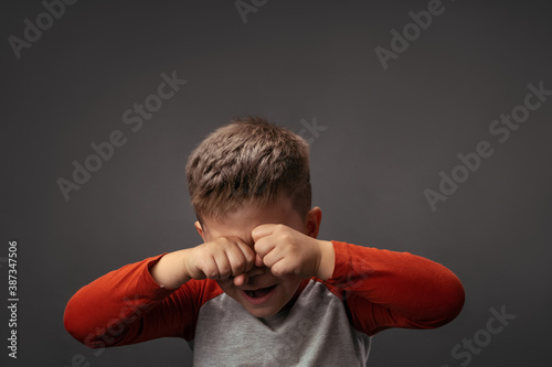 Crying little boy expresses sad emotions. Upset child rubs eyes with fists isolated on gray background. Conflict concept.