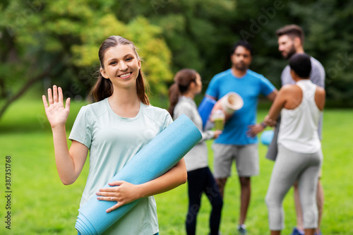 fitness, sport and healthy lifestyle concept - happy smiling young woman with mat waving hand over group of people meeting for yoga class at summer park