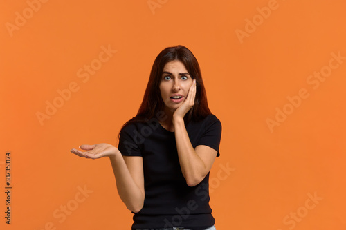 oh no. Shocked caucasian woman wide open eyes in black t-shirt with wondered expression looks at the camera keeps palm raised, and another on cheek, with open mouth isolated on an orange wall