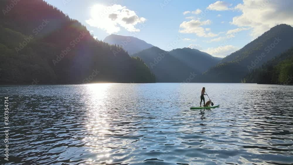 Concept of active tourism and surfing. Aerial drone view of pretty fit woman paddle on sup board at mountain lake during sunset with her dog. Female silhouette doing water sport during summer holidays