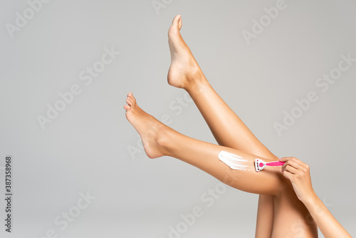 Cropped view of woman with legs in air, shaving leg with safety razor and cream isolated on grey background photo