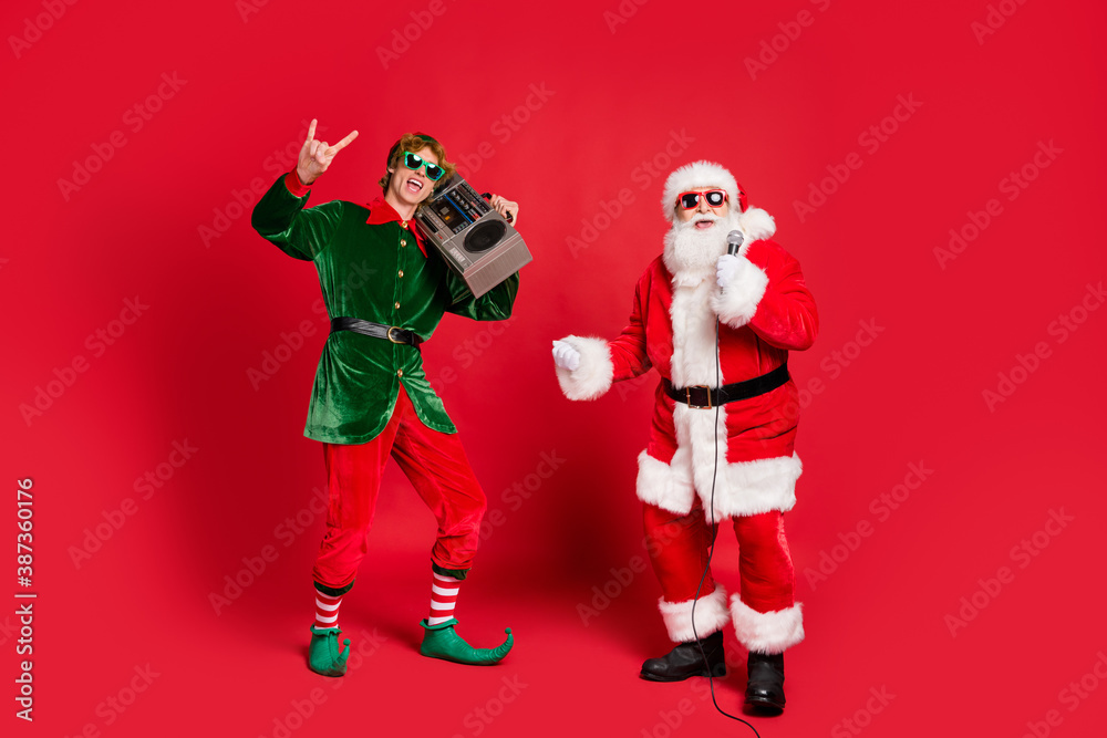 Photo of two crazy men elf hold boom box show horns santa sing mic wear sunglass x-mas costume coat hat isolated red color background Photo of two crazy men elf hold boom box show horns santa sing mic wear sunglass x-mas costume coat hat isolated red color background