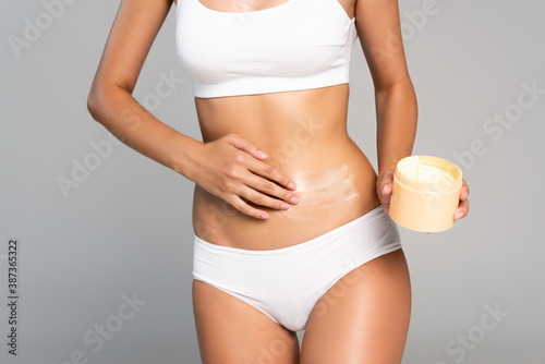 Cropped view of woman in white underwear holding jar and applying cream on belly isolated on grey
