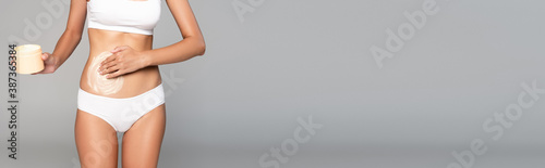 Woman in white underwear holding jar and applying cream on belly on grey, banner