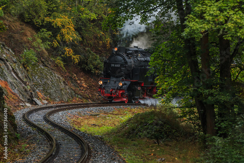 Steam locomotive in a valley in the Harz Mountains. Narrow gauge railway in the mountains in rainy weather in autumn on a track. Trees and rock face along the railroad tracks