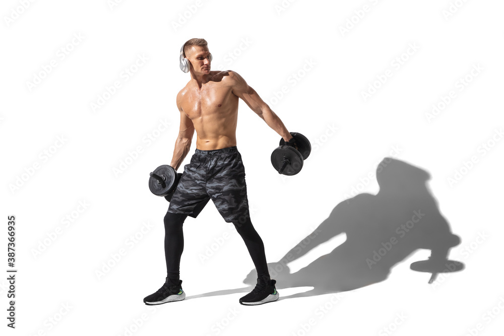 Fototapeta premium Weights and dumbbells. Stylish young male athlete on white studio background, portrait with shadows. Sportive fit model in motion and action. Body building, healthy lifestyle, style concept.