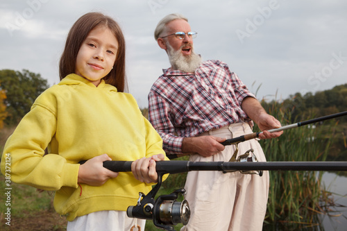 Adorable young girl smiling to the camera, holding fishing rod, enjoying fishing with her grandpa