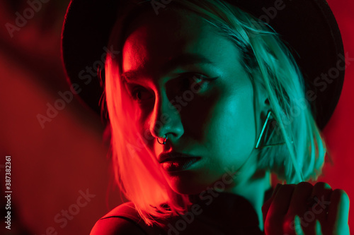 Millennial enigmatic pretty woman with blond dyed hairstyle near glowing neon wall at night. Nose piercing, hipster hat. Mysterious teenager.