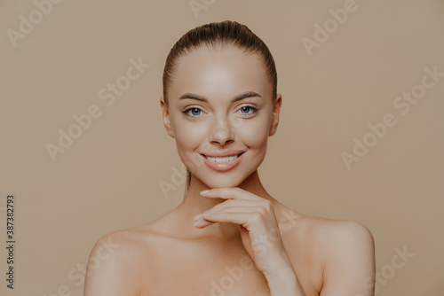 Cheerful young woman with clean glowing skin  keeps hand under chin  smiles gently  has well groomed body  combed hair  poses against beige background. Pure skin  beauty and wellbeing concept