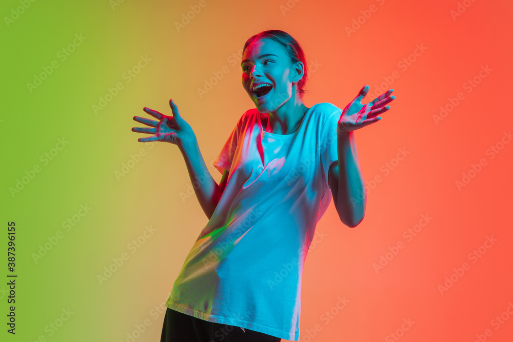 Fototapeta premium Astonished. Young caucasian girl's portrait on gradient green-orange studio background in neon light. Concept of youth, human emotions, facial expression, sales, advertising. Beautiful teen model.