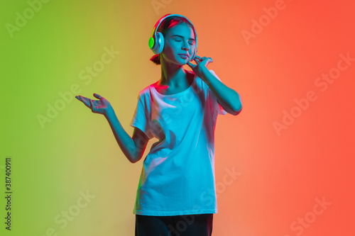 Listening to music with headphones. Young caucasian girl's portrait on gradient green-orange studio background in neon light. Concept of youth, human emotions, facial expression, sales, ad.