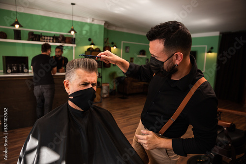 a barber combs an adult male with hand and comb, wears masks pandemic prevention coronavirus, behind two people at the counter talking.