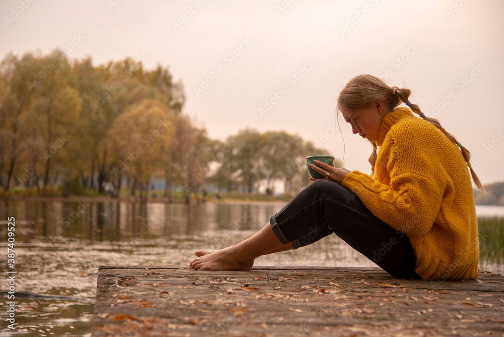 A barefoot girl in a yellow knitted sweater sits on the bridge over the lake and drinks from a green Cup.