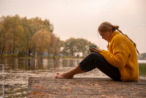 A barefoot girl in a yellow knitted sweater sits on the bridge over the lake and drinks from a green Cup.