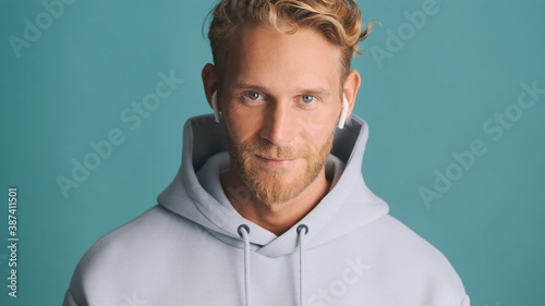 Handsome stylish bearded man in hoodie wear wireless earphones looking confident over colorful background. Modern technology concept © Anton