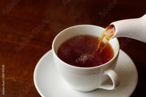 Fotografie Black tea with steam poured into white cup on saucer from a pot.