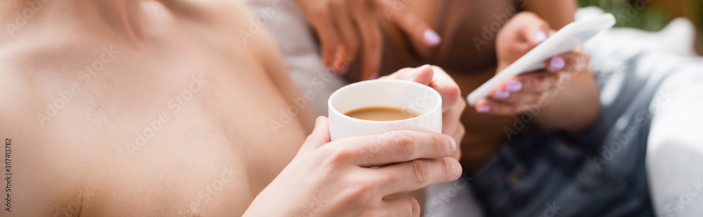 partial view of man holding cup of coffee near girlfriend pointing with finger at smartphone on blurred background, banner partial view of man holding cup of coffee near girlfriend pointing with finger at smartphone on blurred background, banner
