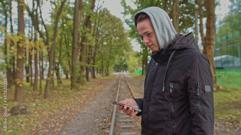 Cropping a view of a millennial hipster, opening and scrolling the phone screen. Young man using a smartphone while walking on the street in the park. A young guy in a hood looks at the phone Cropping a view of a millennial hipster, opening and scrolling the phone screen. Young man using a smartphone while walking on the street in the park. A young guy in a hood looks at the phone