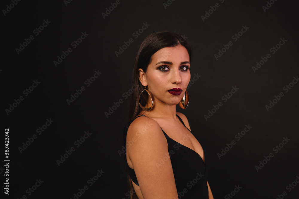 Confident serious woman with long dark hair with bright make up and golden earrings posing over black background Confident serious woman with long dark hair with bright make up and golden earrings posing over black background