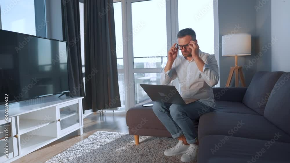Man with glasses sitting on the couch in a cozy room and using laptop. Remote work concept Man with glasses sitting on the couch in a cozy room and using laptop. Remote work concept