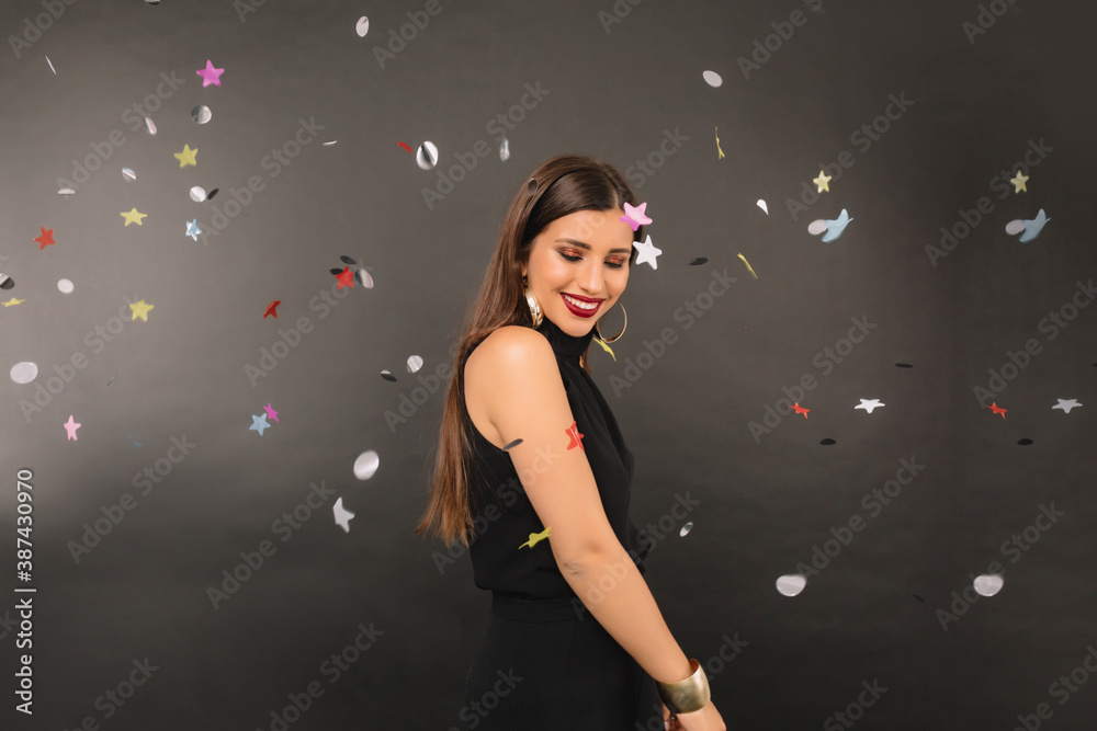 Joyful european woman in black dress celebration under confetti shower and looking at the camera over black background