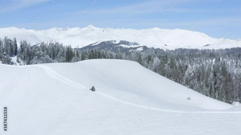People riding on snowmobile with snowboard on snowy highlands at sunny winter day. Snowboarders driving on winter mountain resort. Aerial drone view flying over winter peaks with coniferous fir forest People riding on snowmobile with snowboard on snowy highlands at sunny winter day. Snowboarders driving on winter mountain resort. Aerial drone view flying over winter peaks with coniferous fir forest