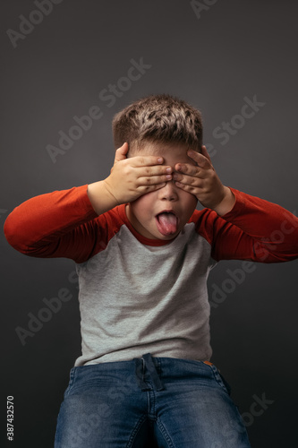 Little boy shows tongue with his eyes closed with hands. Preschooler expresses emotions in studio. © Svyatoslav Lypynskyy