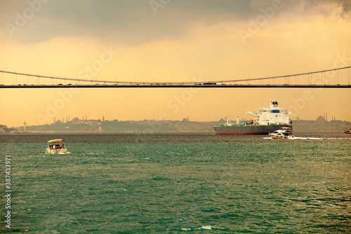 Huge ship is passing under the Bosphorus bridge and a small fishing boat is sailing on a cloudy yellowish day.