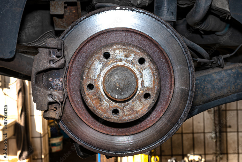 Old and damaged rear brake discs with caliper and brake pads in the car, on a car lift in a workshop.
