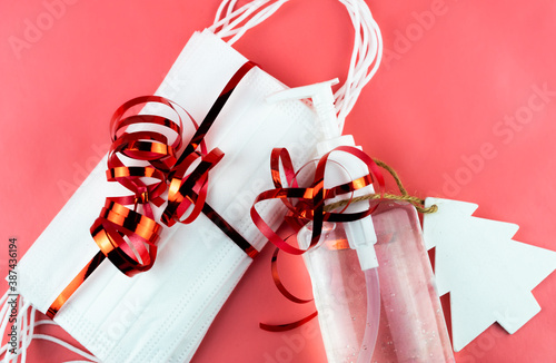 Obraz na płótnie Bottle of hand gel sanitizer and protective masks with Christmas decoration and ribbon as a gift. Celebrating Christmas during coronavirus pandemic lock down and quarantine. Pink background. w salonie