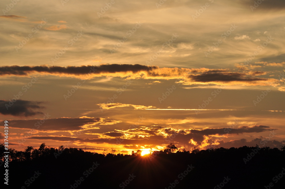 Picture of a sun setting behind a dense forest area followed by mountains.	